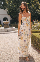 A woman is wearing a long, sleeveless dress with a floral pattern. The dress features a sweetheart neckline with decorative flowers on the straps and a flowy, sheer fabric that reaches the ground. The floral pattern includes purple, yellow, and orange flowers with green leaves on a light, possibly cream or white background. She is also wearing large, round earrings, a bracelet, and white open-toe sandals.